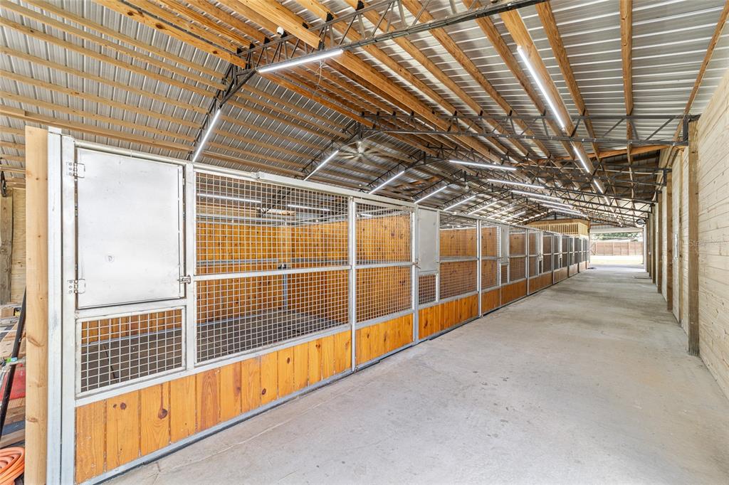A long indoor horse stable aisle features multiple stalls with wood paneling and metal mesh — 1285 NW 102ND A BOULEVARD, WILDWOOD, FL