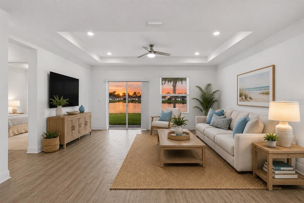 A bright living room features a tray ceiling, wood-look tile floors, and a lake view — 9959 LAST LIGHT GLEN, PARRISH, FL