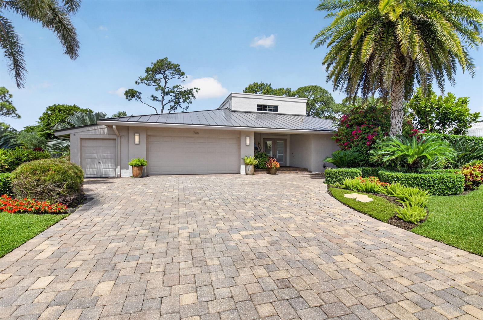 A single-story house with a metal roof, paver driveway, and lush landscaping under a blue sky — 706 Foxpointe Circle, Delray Beach FL 33445, DELRAY BEACH, FL