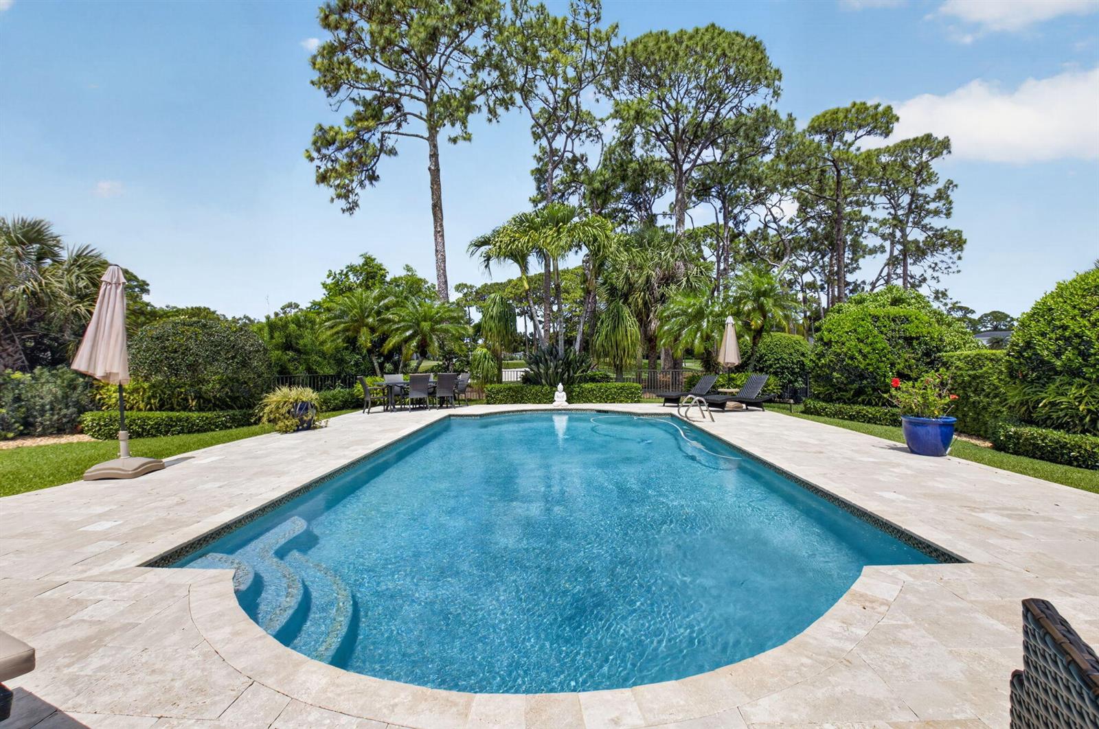 A rectangular swimming pool with stone decking is surrounded by lush landscaping and tall trees — 706 Foxpointe Circle, Delray Beach FL 33445, DELRAY BEACH, FL