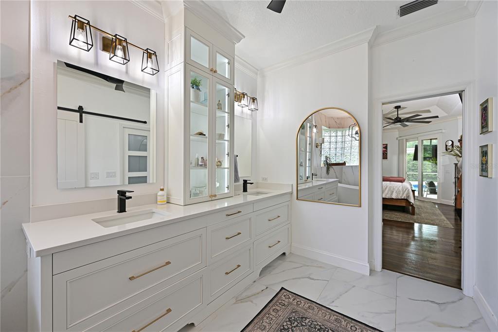 A bright bathroom features a double vanity with white cabinetry, gold hardware, and marble flooring — 5554 MAGNOLIA BLOSSOM LANE, SARASOTA, FL