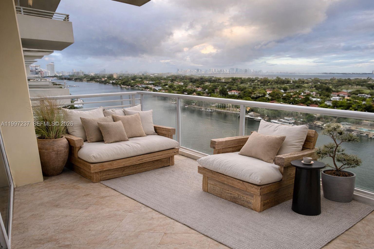 A balcony features wooden lounge furniture, a rug, and potted plants overlooking a waterfront view — 5600 Collins Ave # 16N, Miami Beach FL 33140, MIAMI BEACH, FL