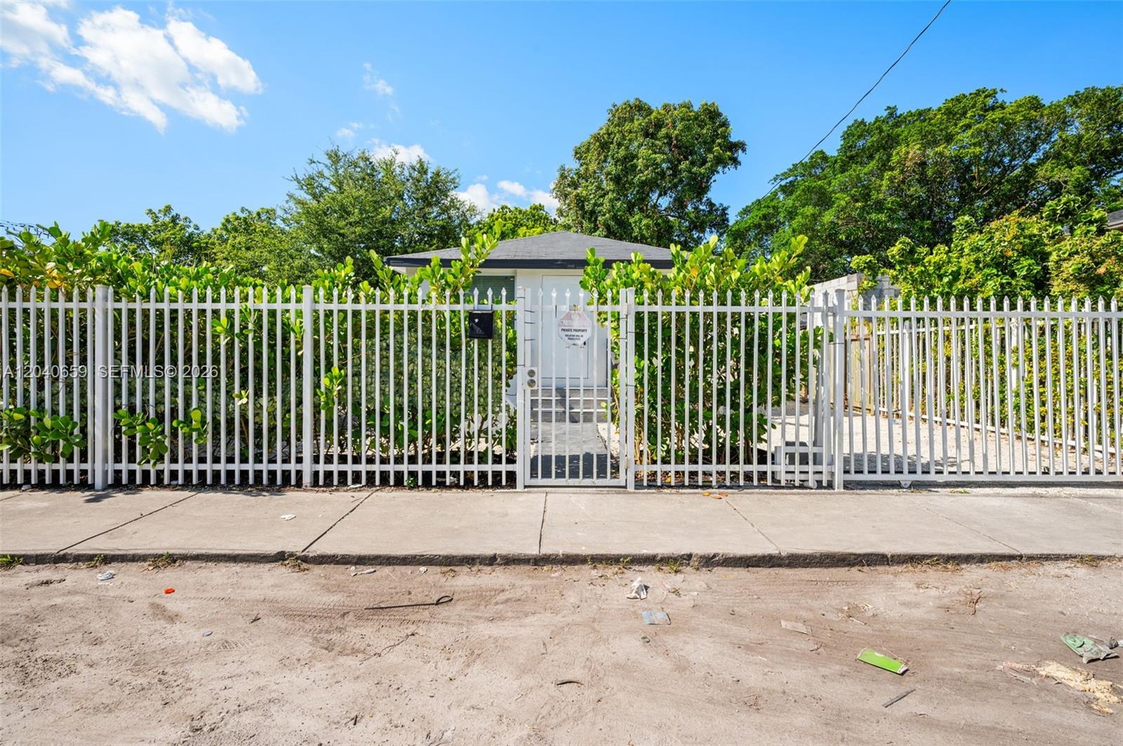 A small house is visible behind a white metal fence and lush green hedges — 1820 NW 63rd St, Miami FL 33147, MIAMI, FL