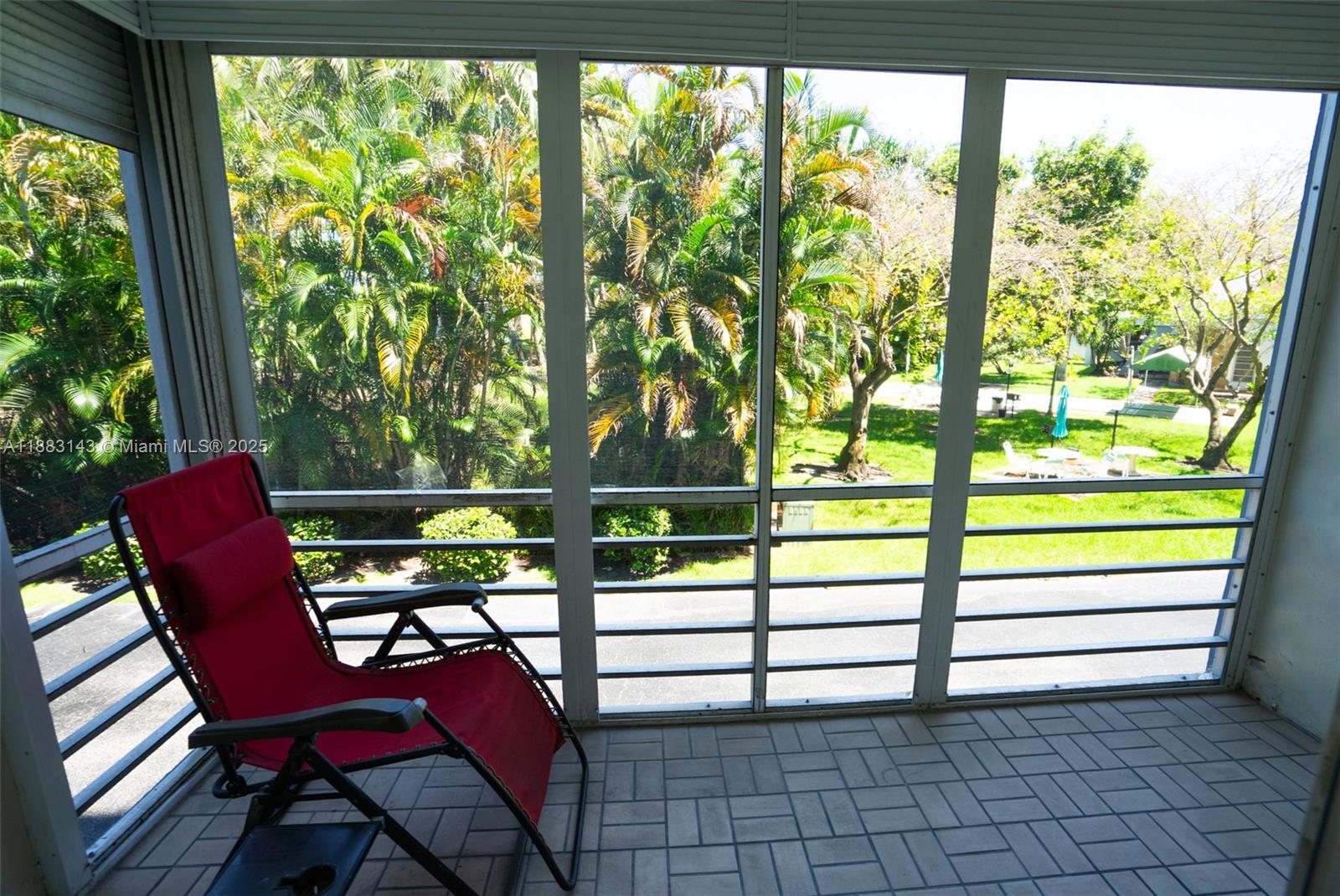 A screened-in balcony features a red lounge chair, tiled flooring, and views of palm trees — 1000 NE 14th Ave # 207, Hallandale Beach FL 33009, HALLANDALE BEACH, FL