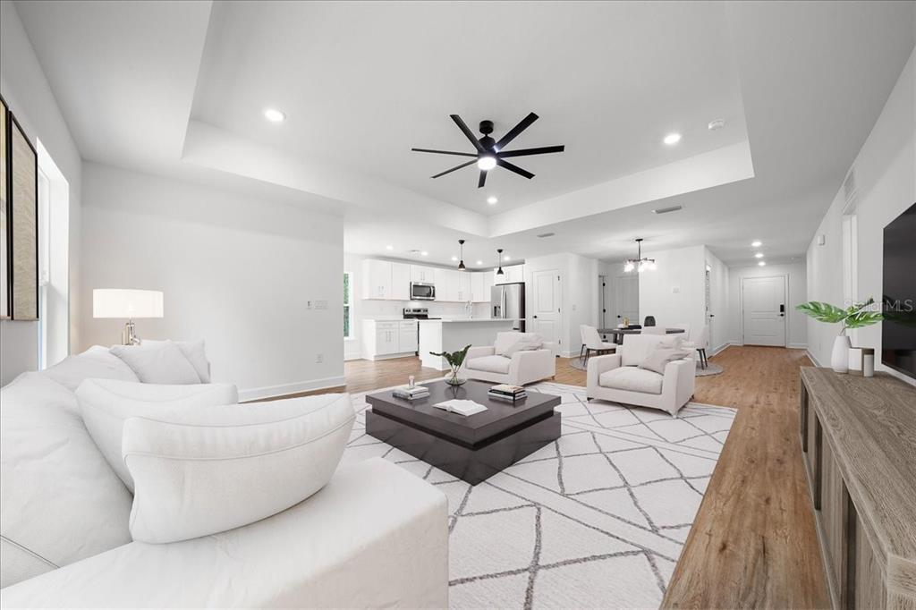 A bright, open-concept living room and kitchen featuring white furniture, wood floors, and tray ceilings — 11041 SPOONBILL ROAD, WEEKI WACHEE, FL