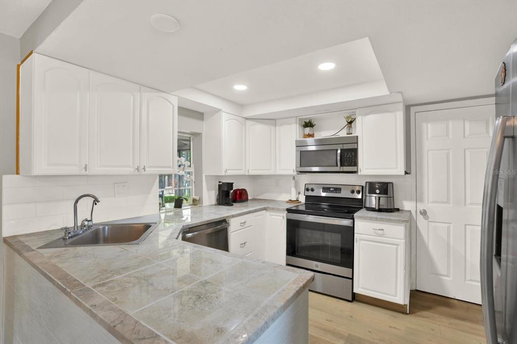 A kitchen features white cabinets, tiled countertops, stainless steel appliances, and a corner sink — 3920 BEACON SQUARE DRIVE, HOLIDAY, FL