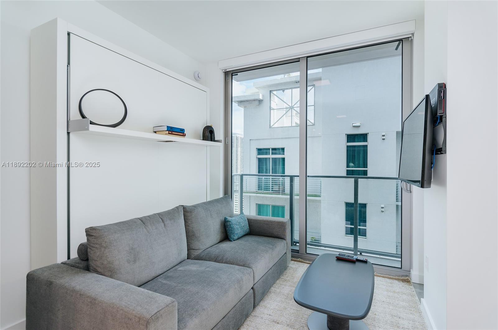 A modern living room features a gray sofa, a wall-mounted bed unit, and balcony access — 227 NE 2nd St # 2508, Miami FL 33132, MIAMI, FL