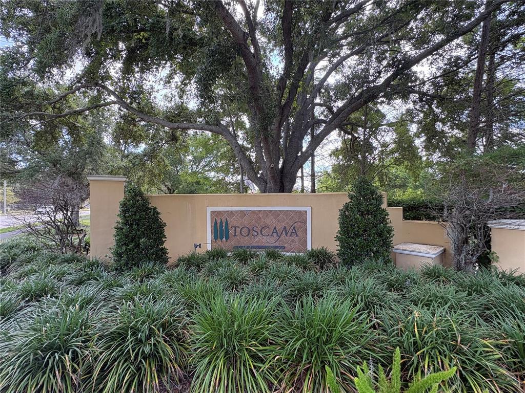 A tan stucco monument sign for Toscana sits behind a landscaped garden under large trees — 6931 LUCCA STREET, ORLANDO, FL