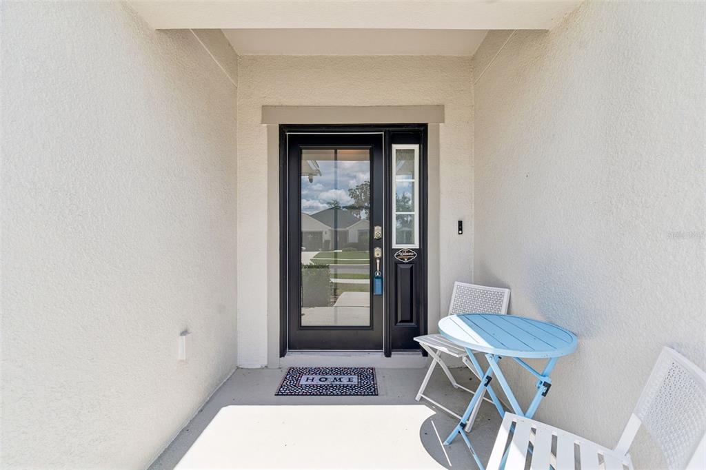 A covered front porch features a black glass-paneled door, a small blue table, and chairs — 6981 TWISTING PINES LOOP, ZEPHYRHILLS, FL