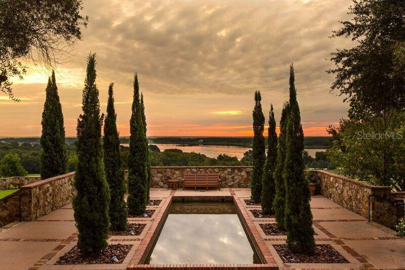 A stone-walled patio features a rectangular reflecting pool, tall cypress trees, and a wooden bench — 15910 VETTA DRIVE, BELLA COLLINA, FL