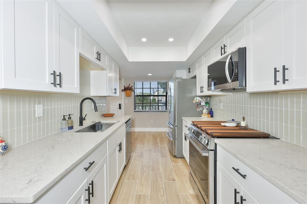 A galley kitchen features white cabinets, light countertops, stainless steel appliances, and wood-look tile flooring — 11800 PARK BOULEVARD UNIT 308, SEMINOLE, FL
