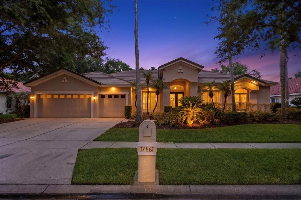 A single-story suburban house with a three-car garage and a landscaped front yard at dusk — 17861 ARBOR GREENE DRIVE, TAMPA, FL