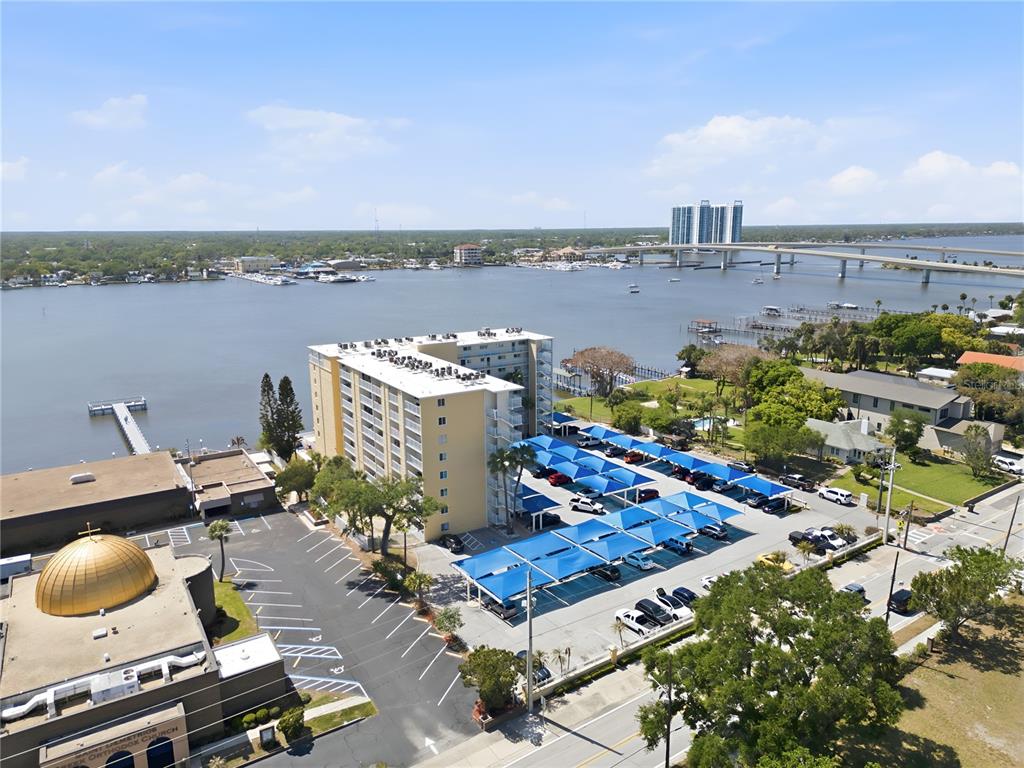 An aerial view shows a multi-story apartment building, a parking lot with shade structures, and water — 145 N HALIFAX AVENUE UNIT 602, DAYTONA BEACH, FL