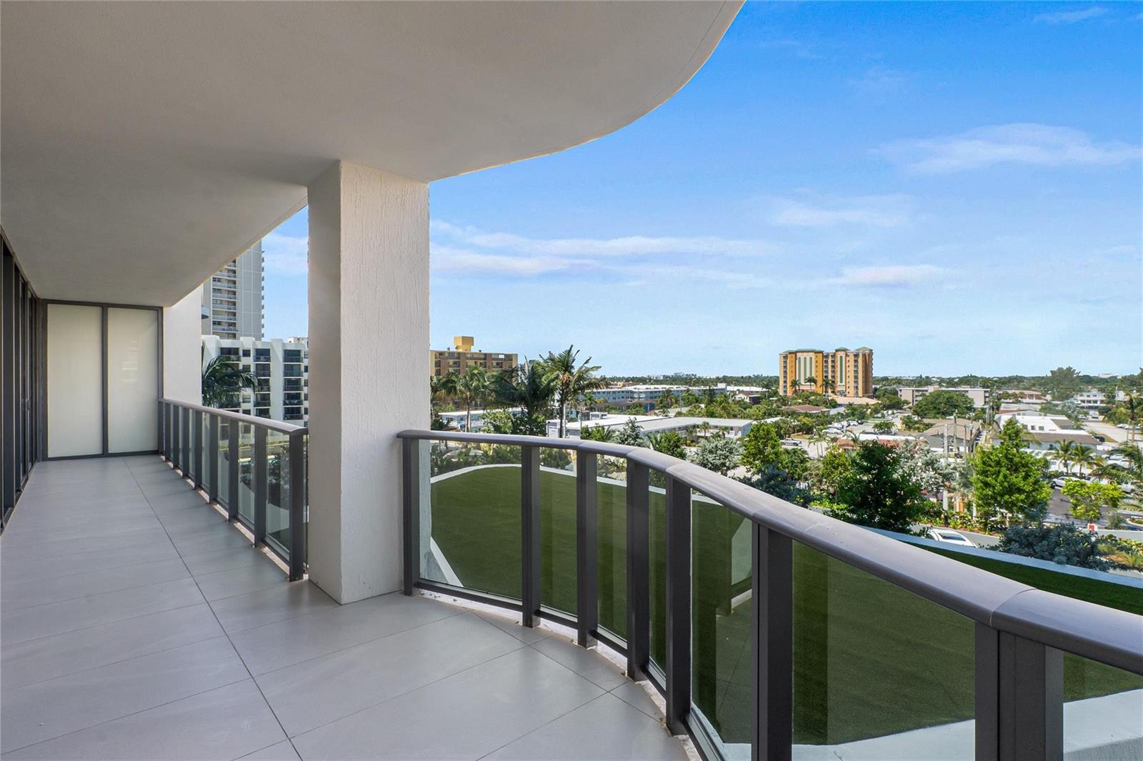 A spacious balcony with gray tile flooring, a glass railing, and views of surrounding buildings — 900 N Ocean Boulevard # 504, Pompano Beach FL 33062, POMPANO BEACH, FL
