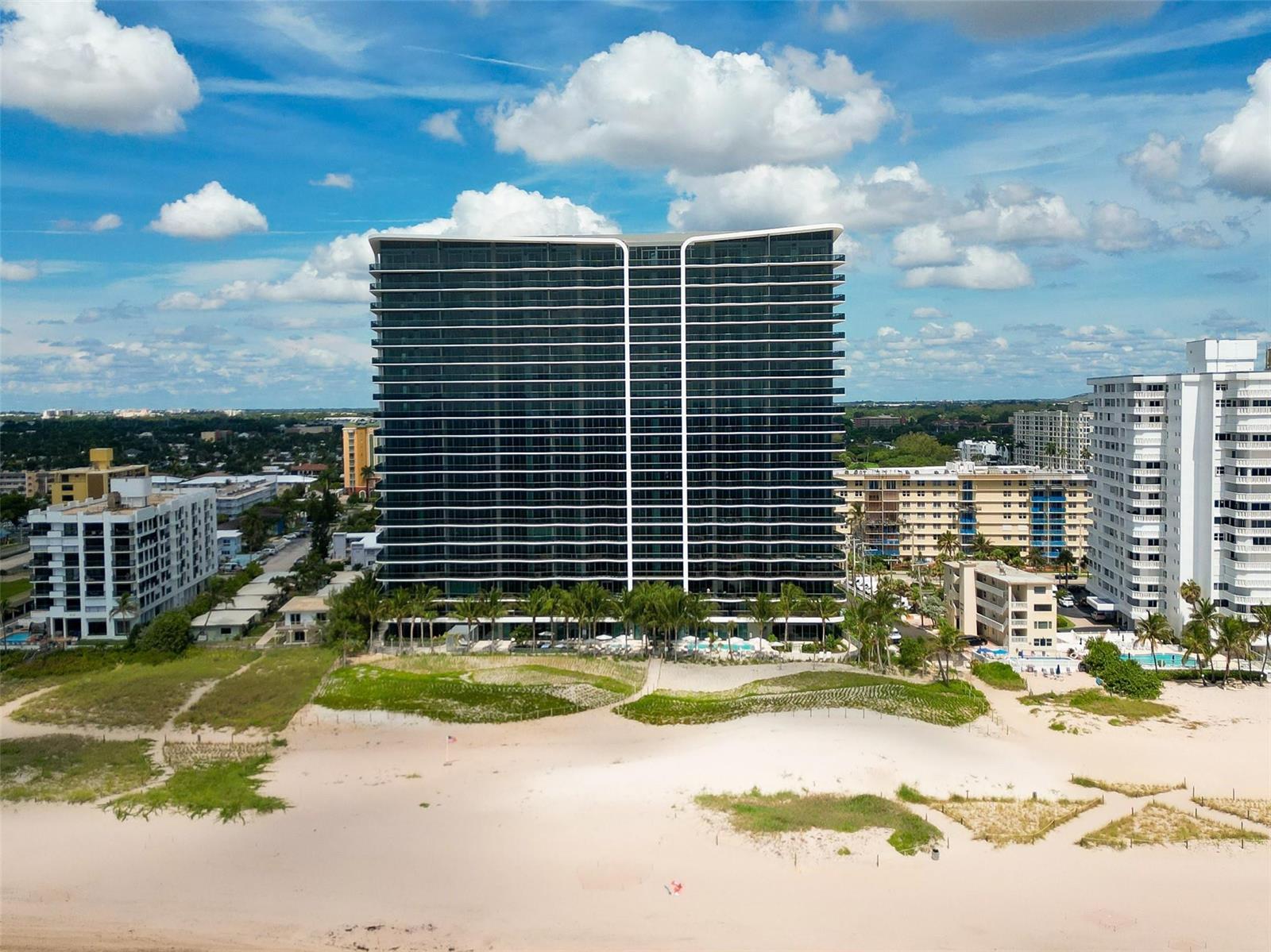 An aerial view of a tall, modern beachfront condominium building surrounded by sand and palm trees — 900 N Ocean Boulevard # 504, Pompano Beach FL 33062, POMPANO BEACH, FL