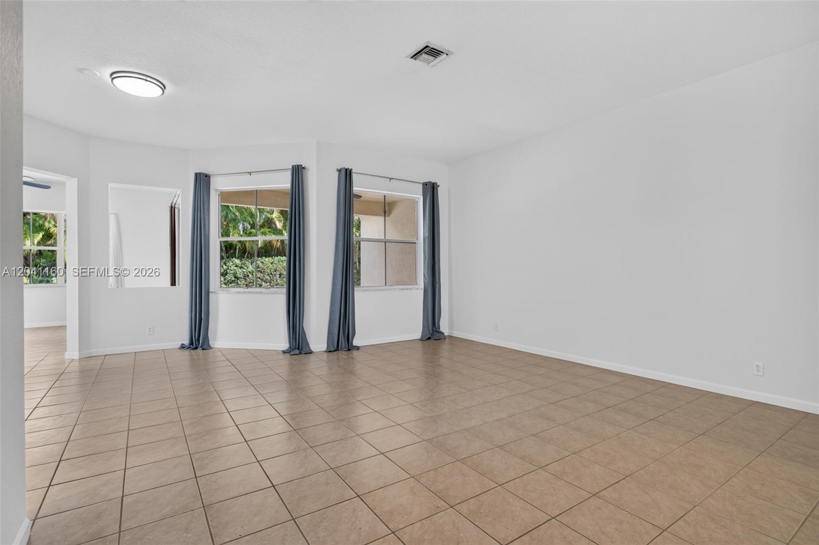 An empty room with beige tile flooring, white walls, and two windows with blue curtains — 786 Nandina Dr, Weston FL 33327, WESTON, FL