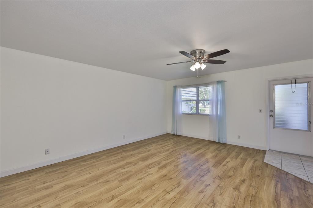 An empty living room features light wood-look flooring, white walls, a ceiling fan, and a door — 1811 BEDFORD LANE UNIT 153, SUN CITY CENTER, FL