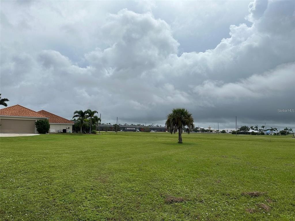 A large, grassy vacant lot sits under a cloudy sky next to a residential house — 24472 MANIZALES COURT, PUNTA GORDA, FL