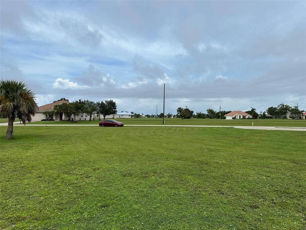 A wide, grassy vacant lot sits in front of a paved road and suburban houses — 24472 MANIZALES COURT, PUNTA GORDA, FL