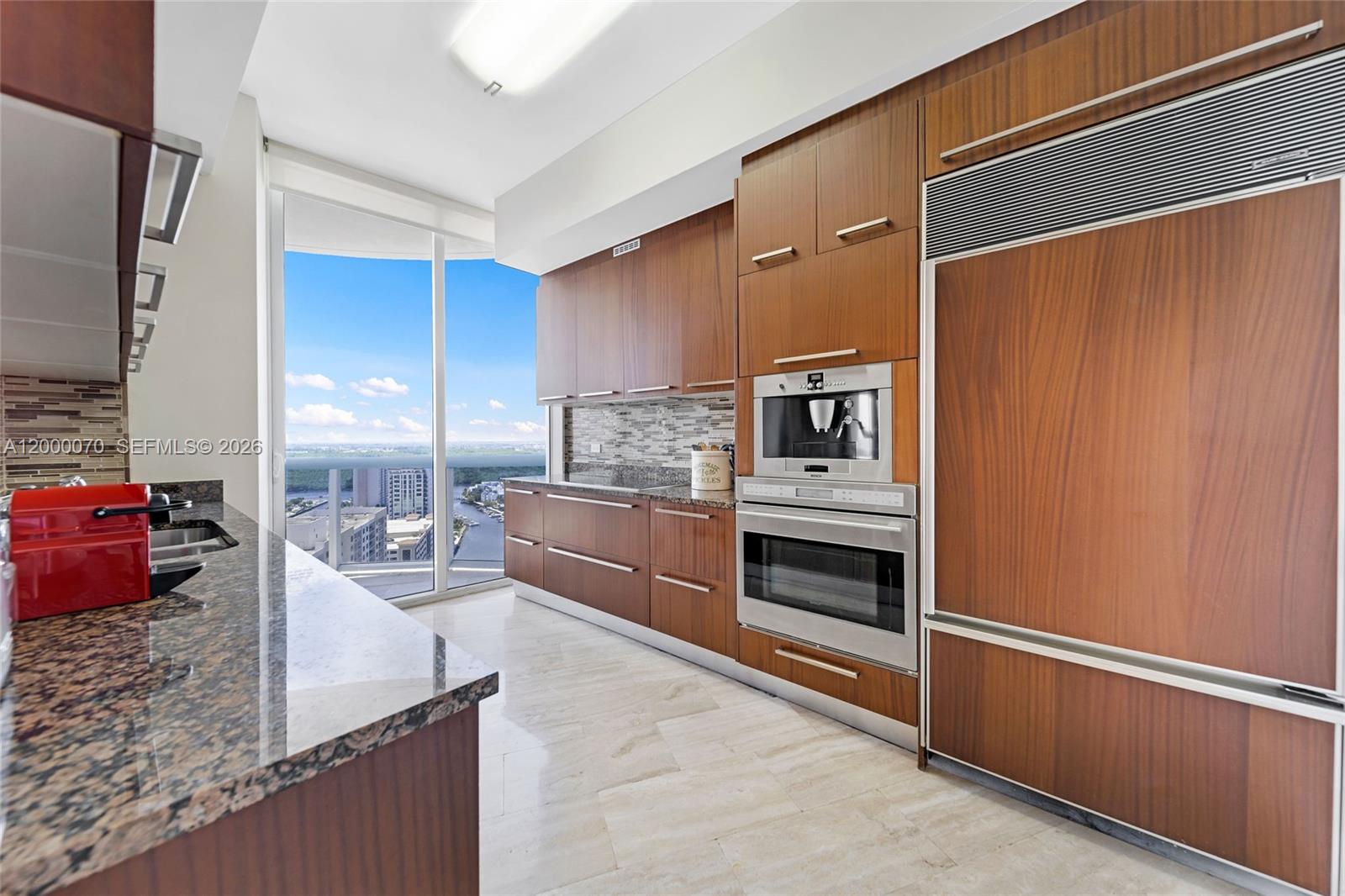 A modern kitchen features wood cabinetry, stainless steel appliances, granite countertops, and floor-to-ceiling windows — SUNNY ISLES BEACH, FL