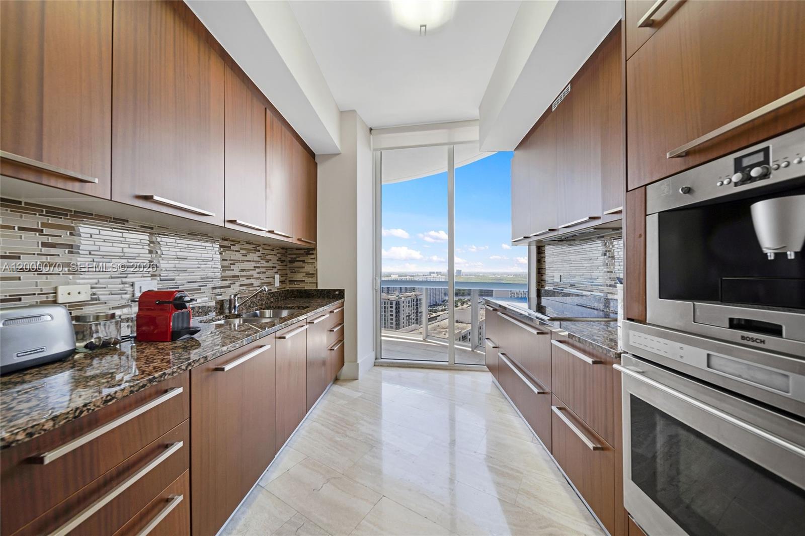 A galley kitchen features wood cabinetry, granite countertops, a built-in coffee maker, and balcony access — SUNNY ISLES BEACH, FL
