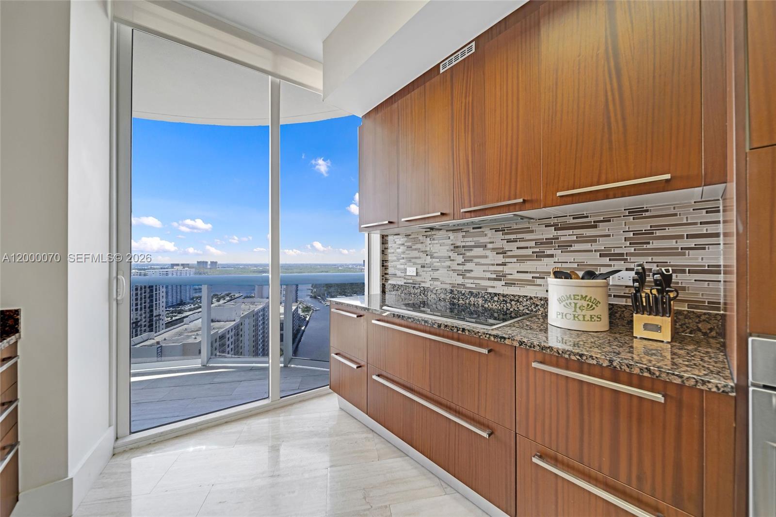 A modern kitchen features wood cabinets, granite countertops, and a balcony with a city view — SUNNY ISLES BEACH, FL