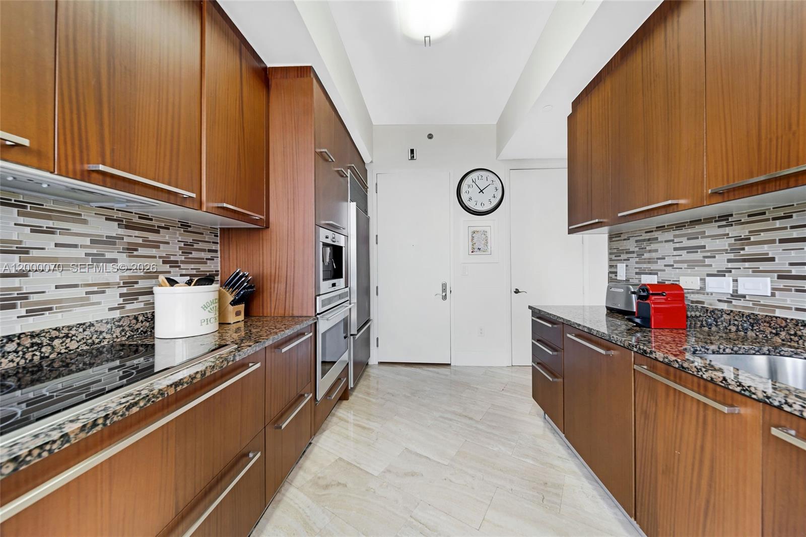A galley kitchen features wood cabinets, granite countertops, a tiled backsplash, and light stone flooring — SUNNY ISLES BEACH, FL