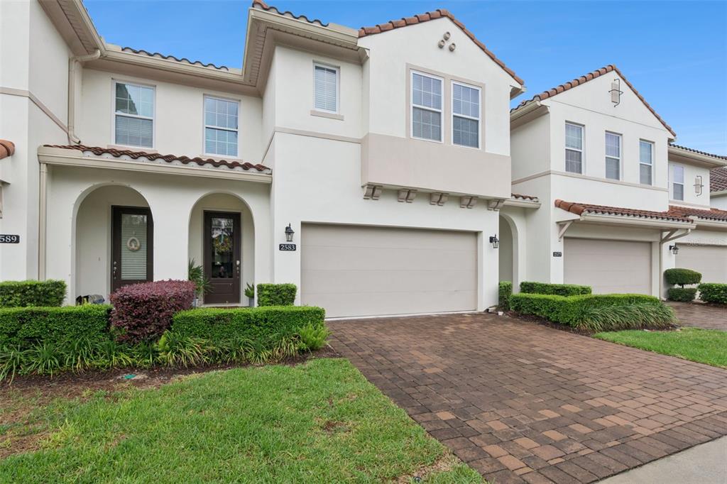 A two-story townhouse features a beige stucco exterior, a two-car garage, and landscaped hedges — 2583 CIRCOLO LANE, APOPKA, FL