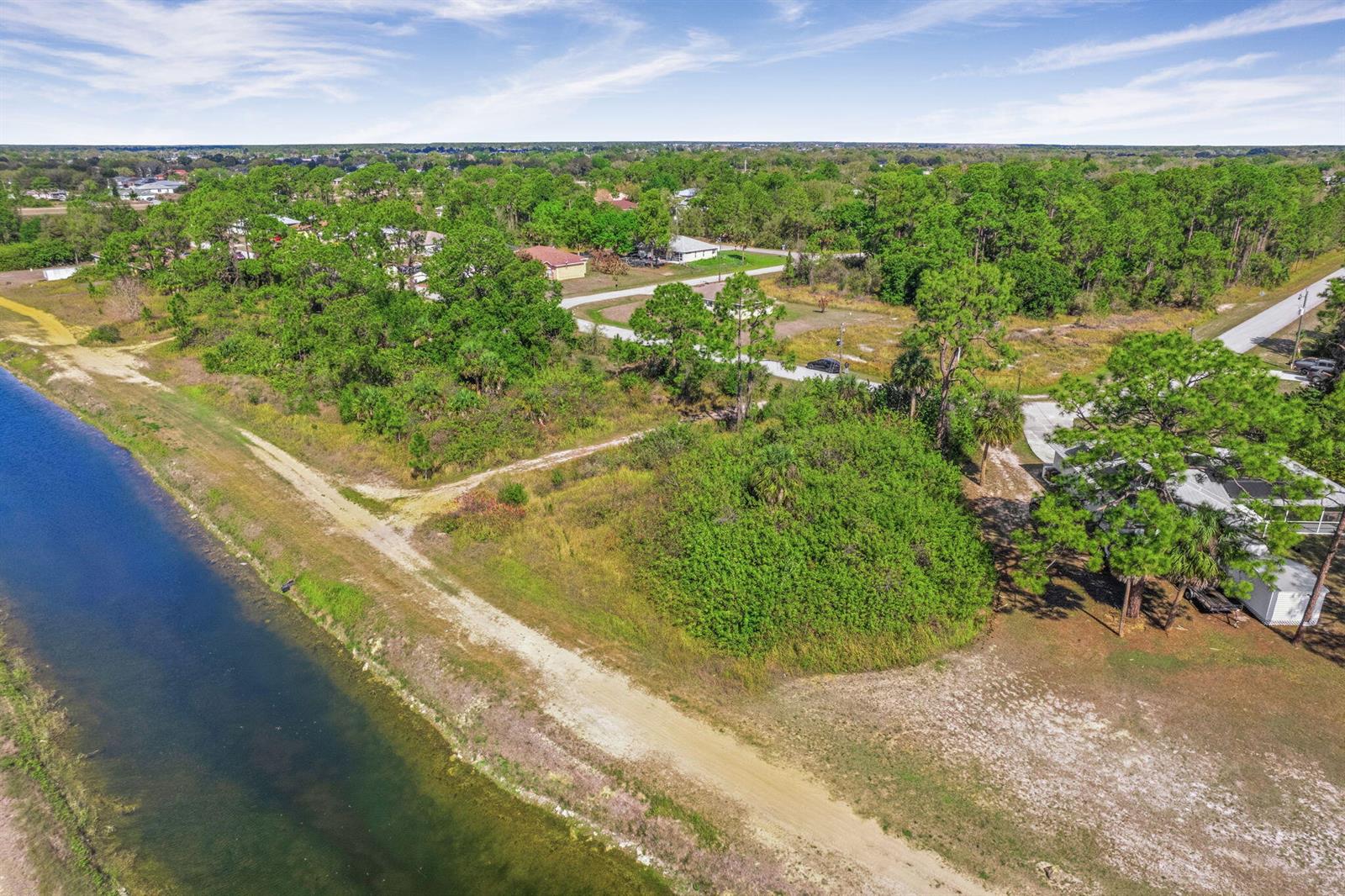 An aerial view shows a dirt path running alongside a canal near wooded residential lots — 755 Longbow Lane, Lehigh Acres FL 33972, LEHIGH ACRES, FL