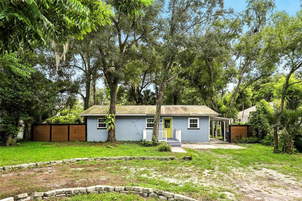 A single-story blue house with a yellow front door sits among many large trees — 1412 E VOORHIS AVENUE, DELAND, FL