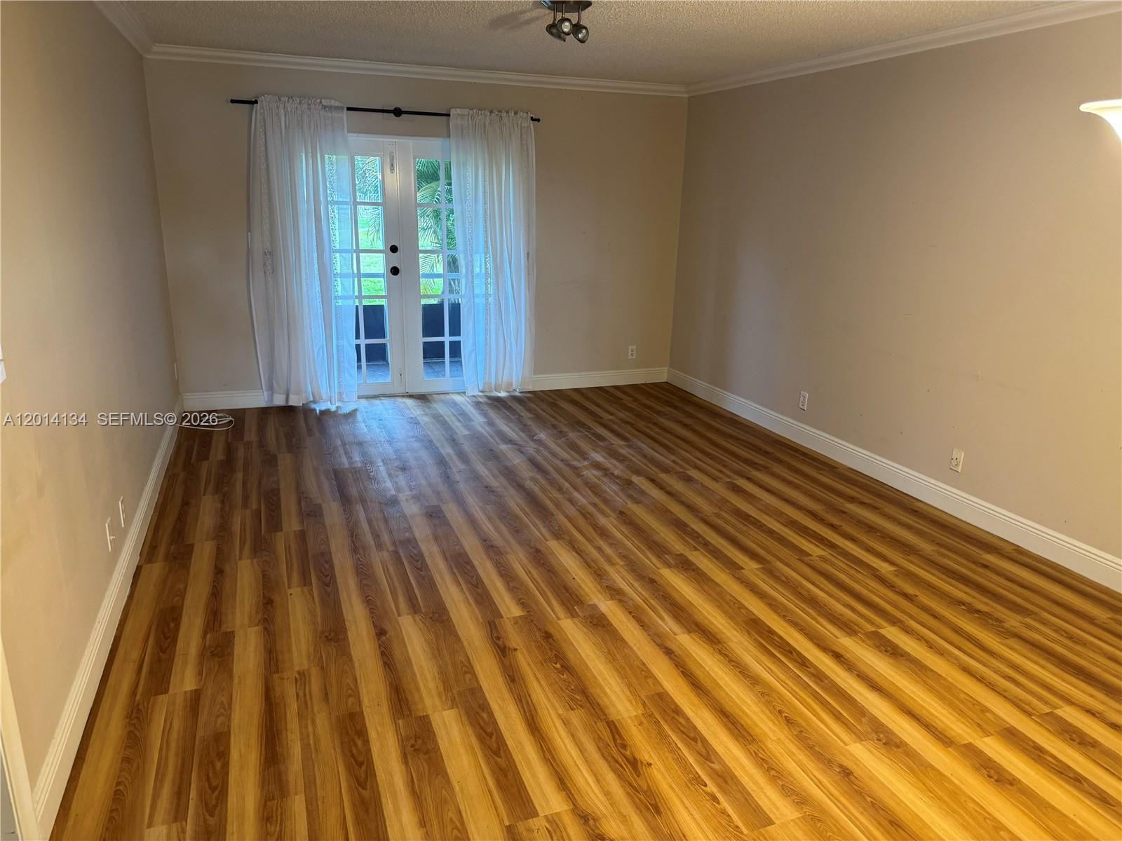 An empty room features wood-look flooring, beige walls, and white French doors with sheer curtains — 3774 Inverrary Blvd # 106P, Lauderhill FL 33319, LAUDERHILL, FL