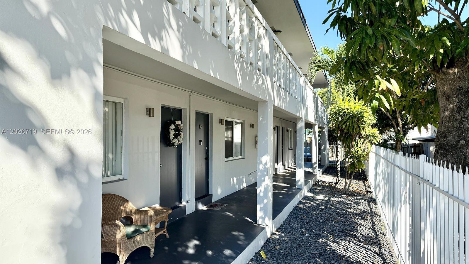 A white two-story apartment building exterior features a covered walkway, wicker chair, and gravel ground — 42 SW 14th St # 103, Dania Beach FL 33004, DANIA BEACH, FL