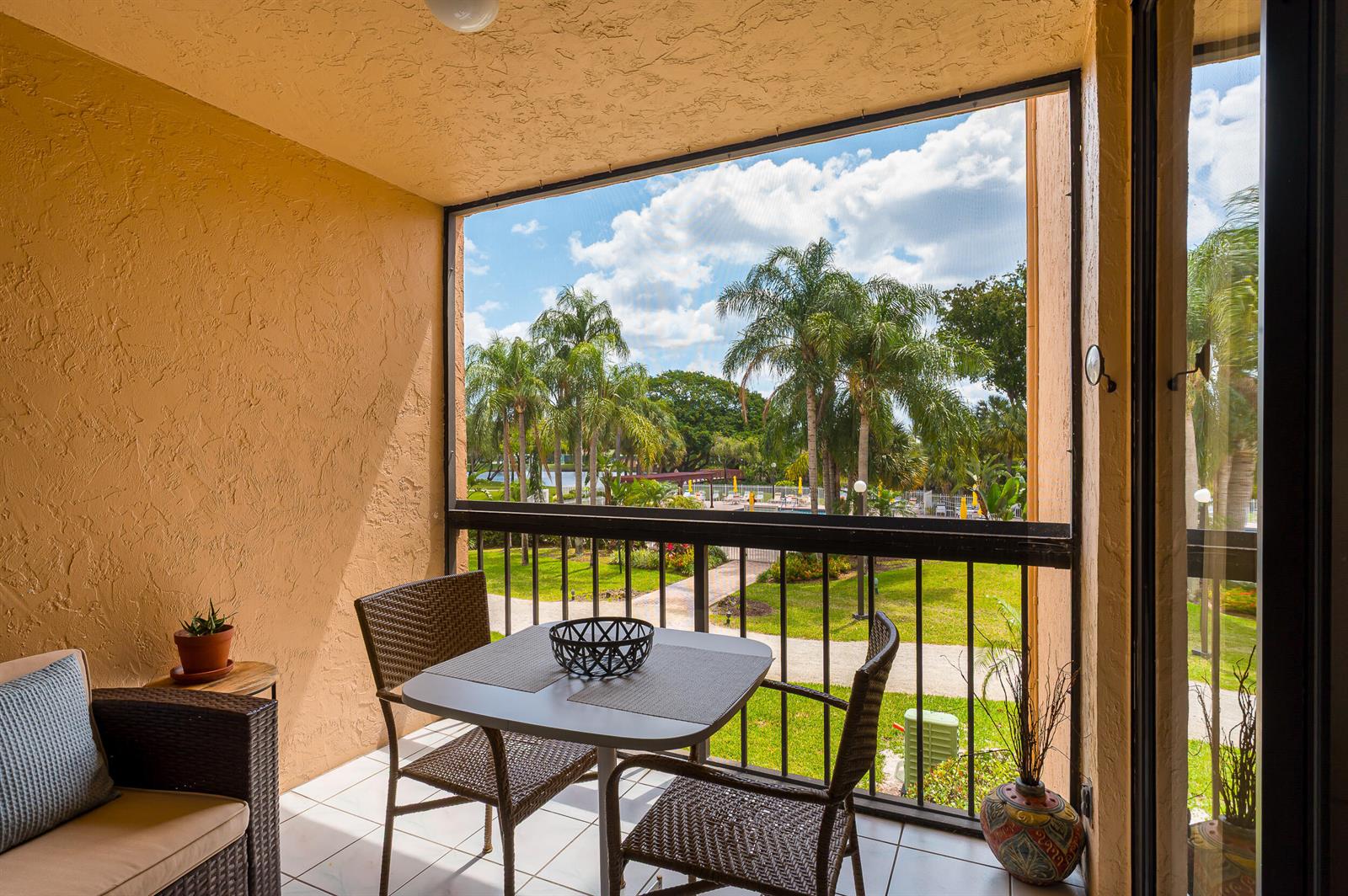 A covered balcony with wicker furniture overlooks a grassy courtyard with palm trees and a pool — 2255 Lindell Boulevard # 4207, Delray Beach FL 33444, DELRAY BEACH, FL