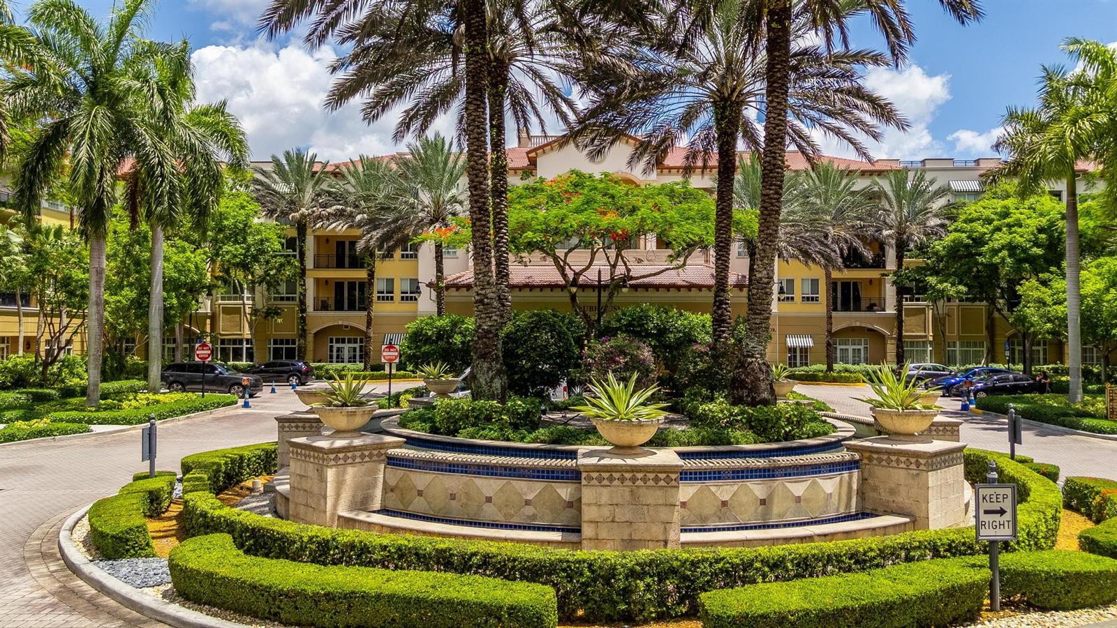 A circular driveway with a central fountain and landscaping in front of a large building — 16102 Emerald Estates Drive # 128, Weston FL 33331, WESTON, FL