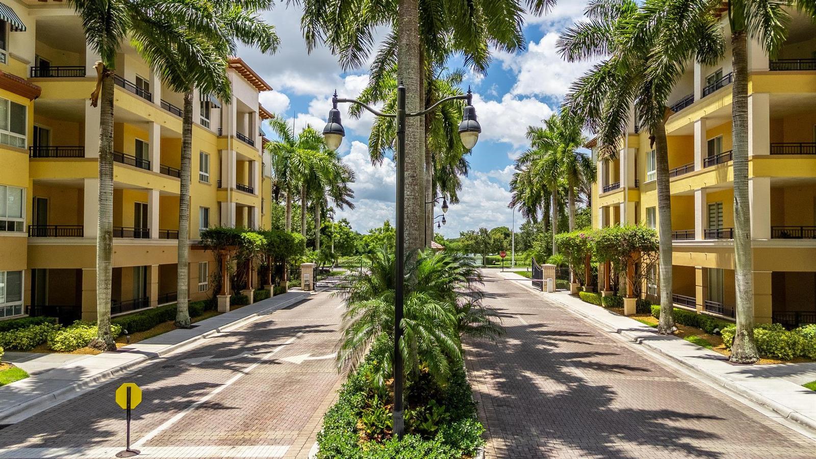 A paved driveway leads between two yellow apartment buildings lined with palm trees — 16102 Emerald Estates Drive # 128, Weston FL 33331, WESTON, FL