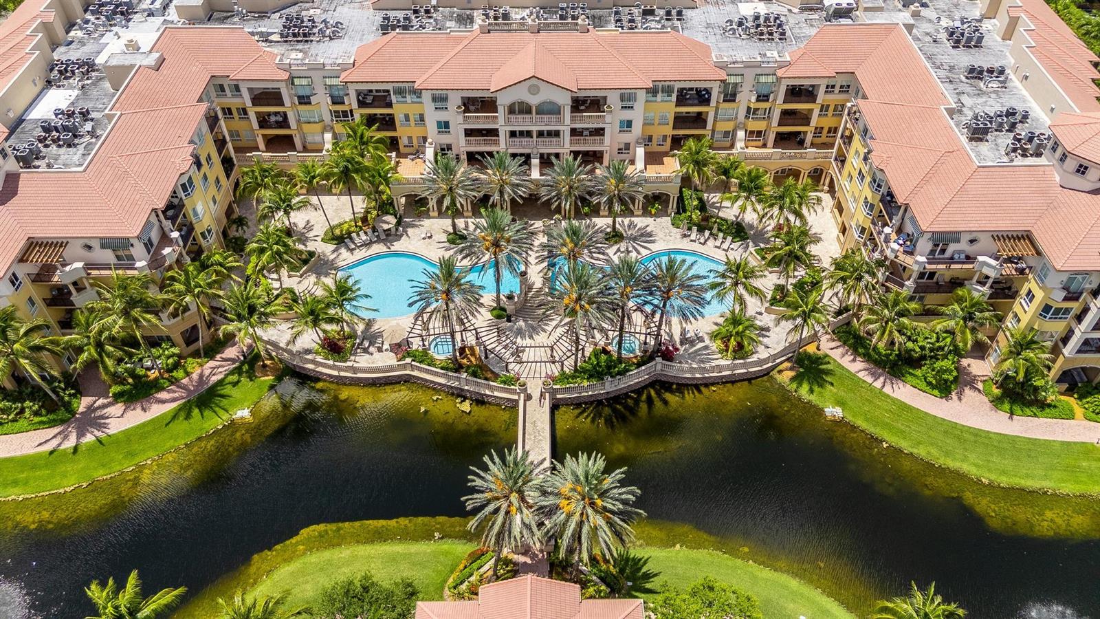 An aerial view of a resort-style apartment complex featuring a central pool and a pond — 16102 Emerald Estates Drive # 128, Weston FL 33331, WESTON, FL