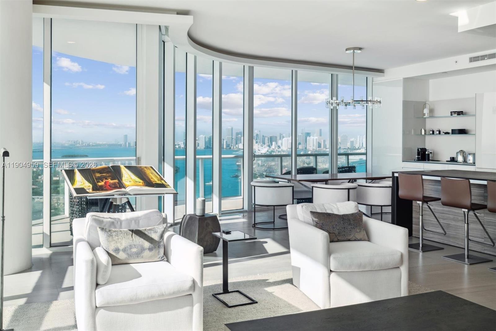 A modern living room features white armchairs, a kitchen island, and floor-to-ceiling windows overlooking water — 100 S Pointe Dr # 3203, Miami Beach FL 33139, MIAMI BEACH, FL
