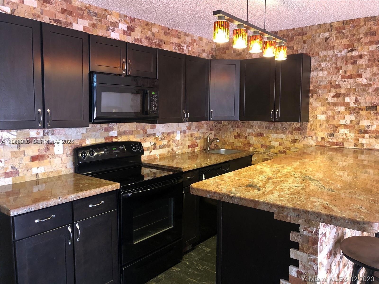 A kitchen features dark cabinets, granite countertops, and a textured stone tile backsplash — 600 SE Three Islands Blvd # 1018, Hallandale Beach FL 33009, HALLANDALE BEACH, FL