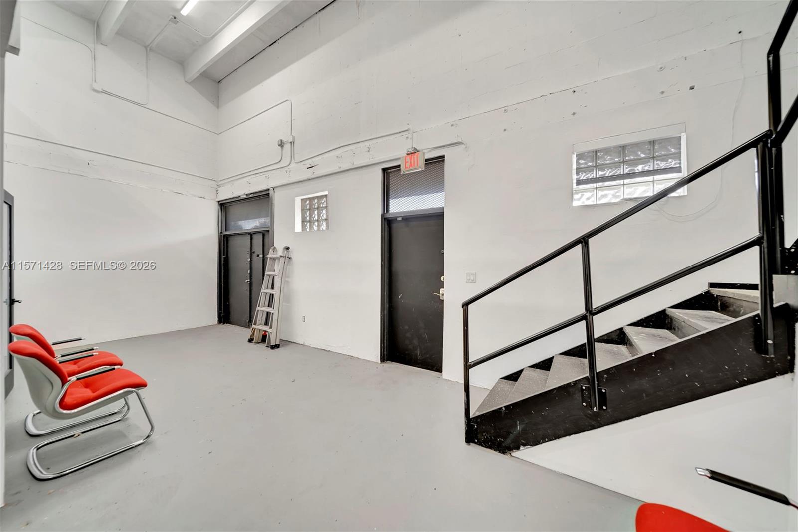 An industrial-style room features white walls, a gray floor, red chairs, and a metal staircase — 67 NW 166th St, Miami FL 33169, MIAMI, FL