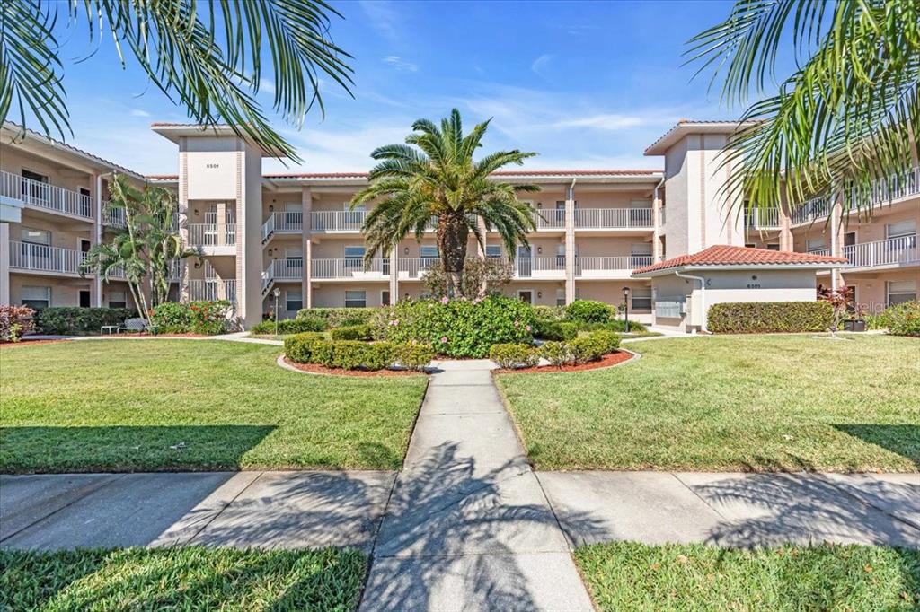A multi-story apartment building features a central palm tree and a paved walkway — 6501 STONE RIVER ROAD UNIT 209, BRADENTON, FL