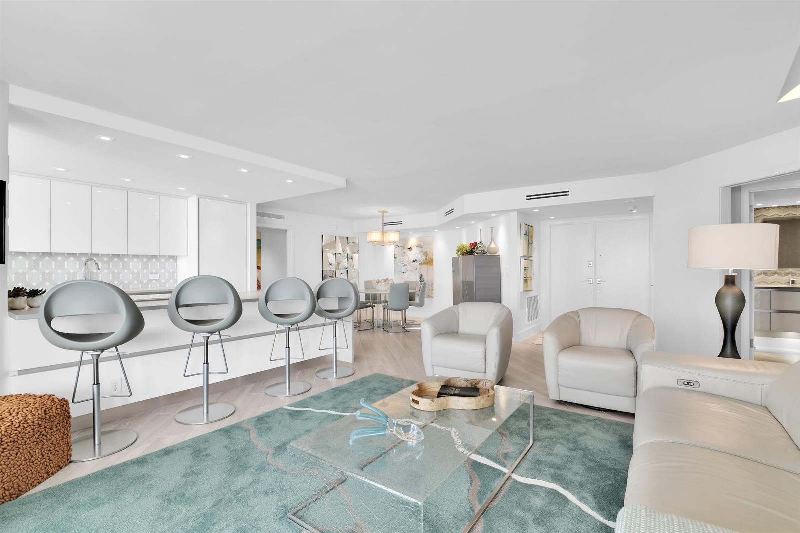 A modern open-concept living room and kitchen featuring white cabinetry, bar stools, and teal rug — 550 S Ocean Boulevard # 1004, Boca Raton FL 33432, BOCA RATON, FL