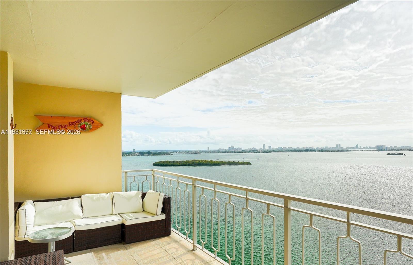 A balcony with outdoor seating overlooks a large body of water and a distant city skyline — 11111 Biscayne Blvd # 20E, Miami FL 33181, MIAMI, FL