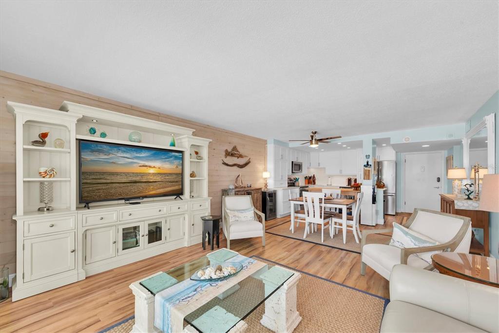 A living room with a large entertainment center, wood-look flooring, and an open kitchen area — 420 64TH AVENUE UNIT 402E, ST PETE BEACH, FL