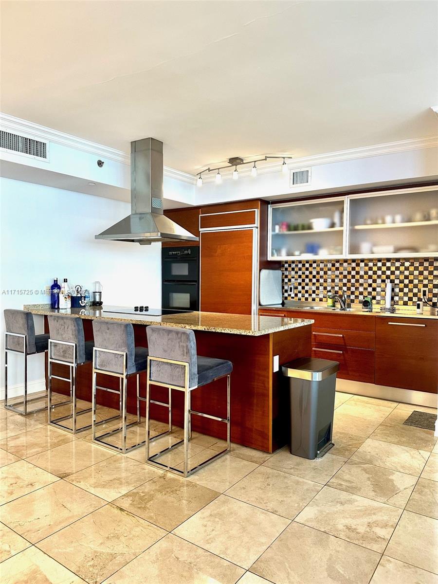A modern kitchen features wood cabinetry, a granite island with four barstools, and tile flooring — 1850 S Ocean Dr # 3310, Hallandale Beach FL 33009, HALLANDALE BEACH, FL