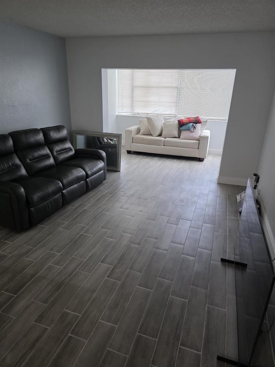 A living room features gray wood-look tile flooring, a black leather sofa, and a white sofa — 3130 Holiday Springs Boulevard # 104, Margate FL 33063, MARGATE, FL