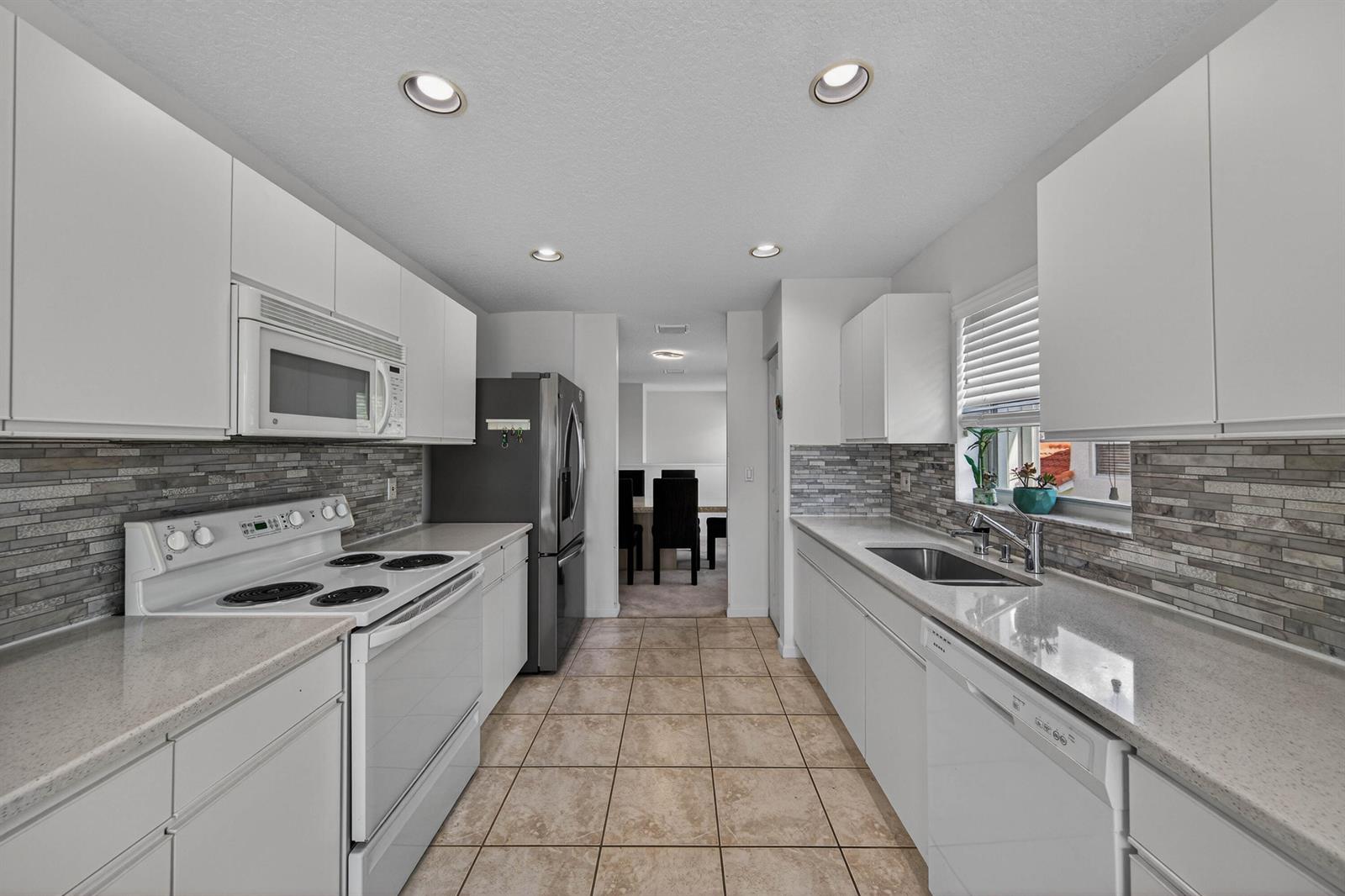 A galley kitchen features white cabinets, stone tile backsplash, light countertops, and stainless steel appliances — 7676 Springwater Place # 201, Boynton Beach FL 33437, BOYNTON BEACH, FL