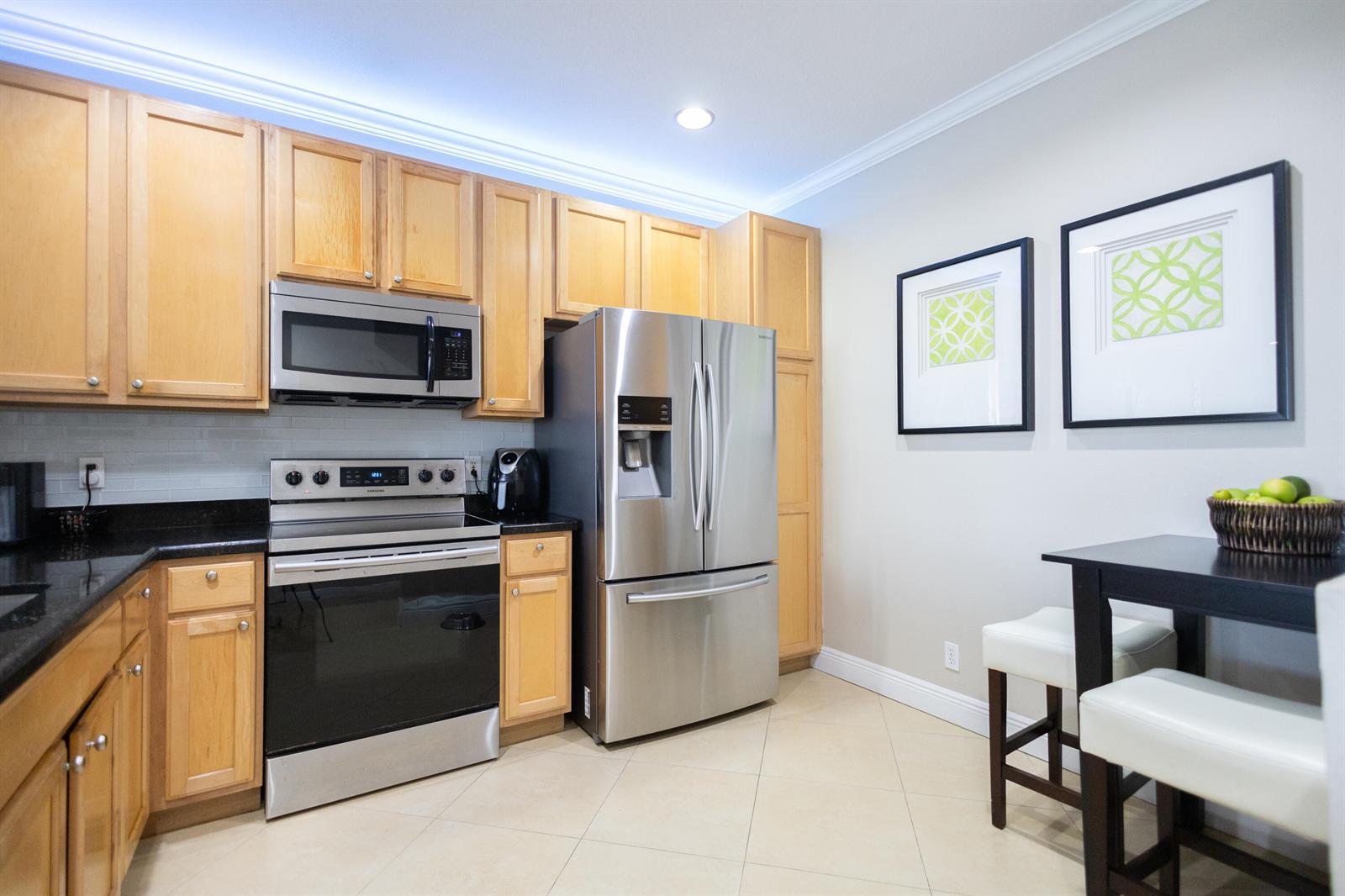 A kitchen features light wood cabinets, stainless steel appliances, black countertops, and a small dining area — 8115 Bellagio Lane, Boynton Beach FL 33472, BOYNTON BEACH, FL