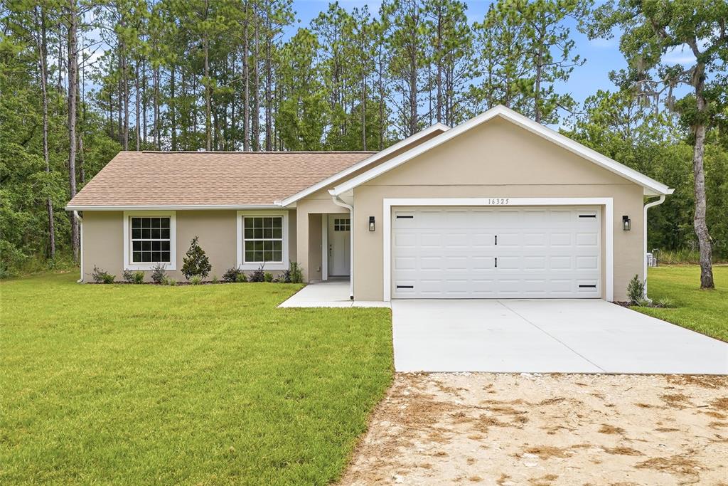 A single-story house with a beige stucco exterior, white garage door, and surrounding pine trees — 16325 FORCE ROAD, BROOKSVILLE, FL