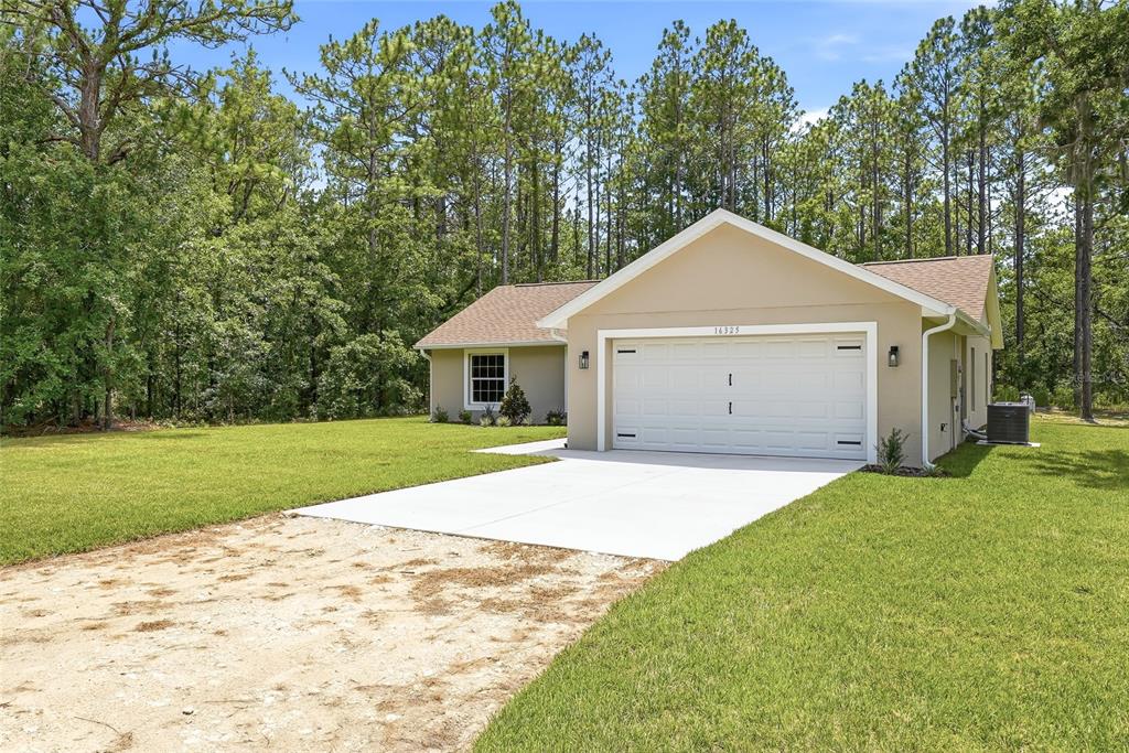 A tan single-story house with a two-car garage sits on a grassy lot near trees — 16325 FORCE ROAD, BROOKSVILLE, FL