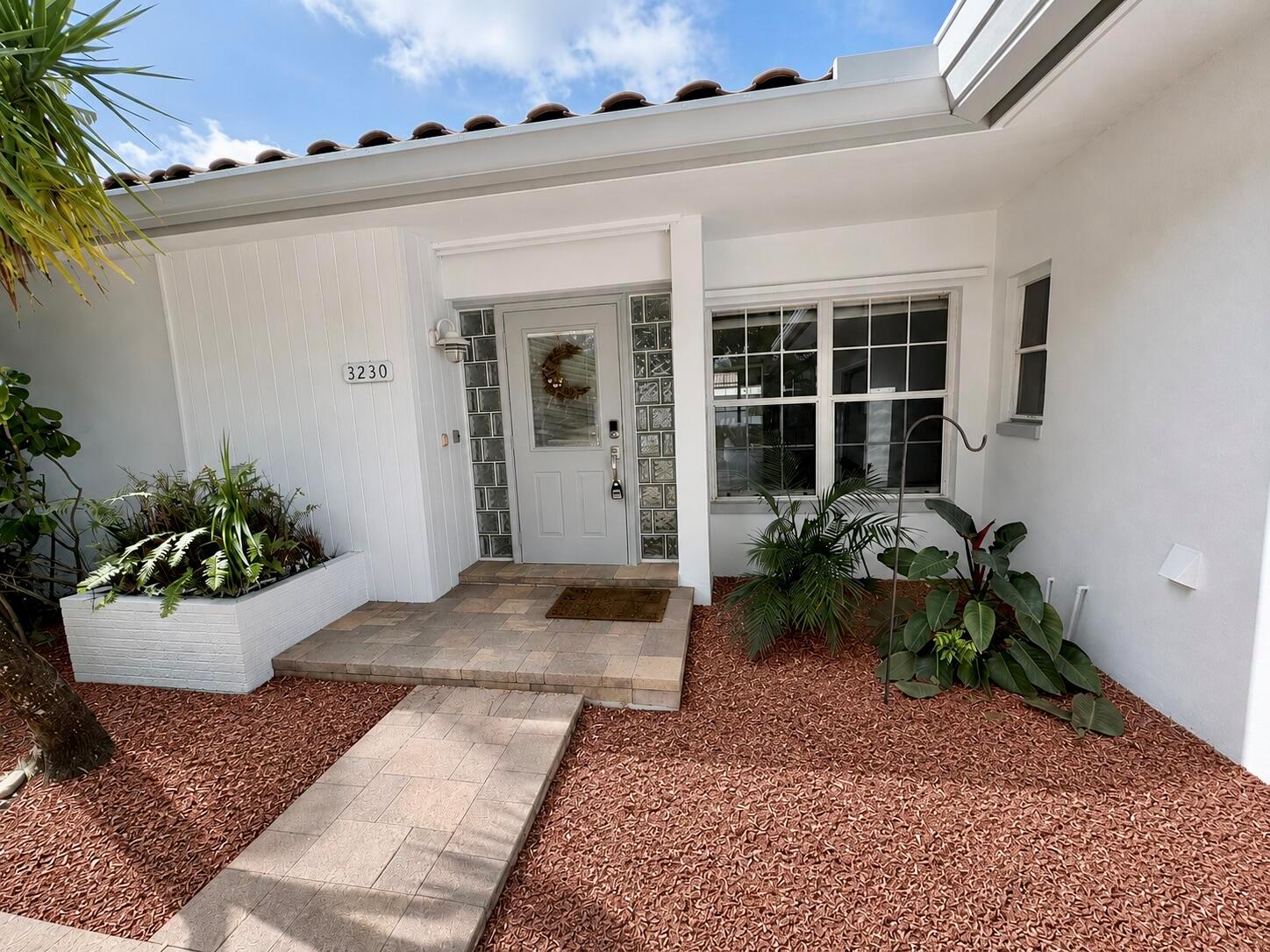 A white house exterior features a paved walkway, glass block accents, and red mulch landscaping — 3230 Aqua Vista Drive, Pompano Beach FL 33062, POMPANO BEACH, FL